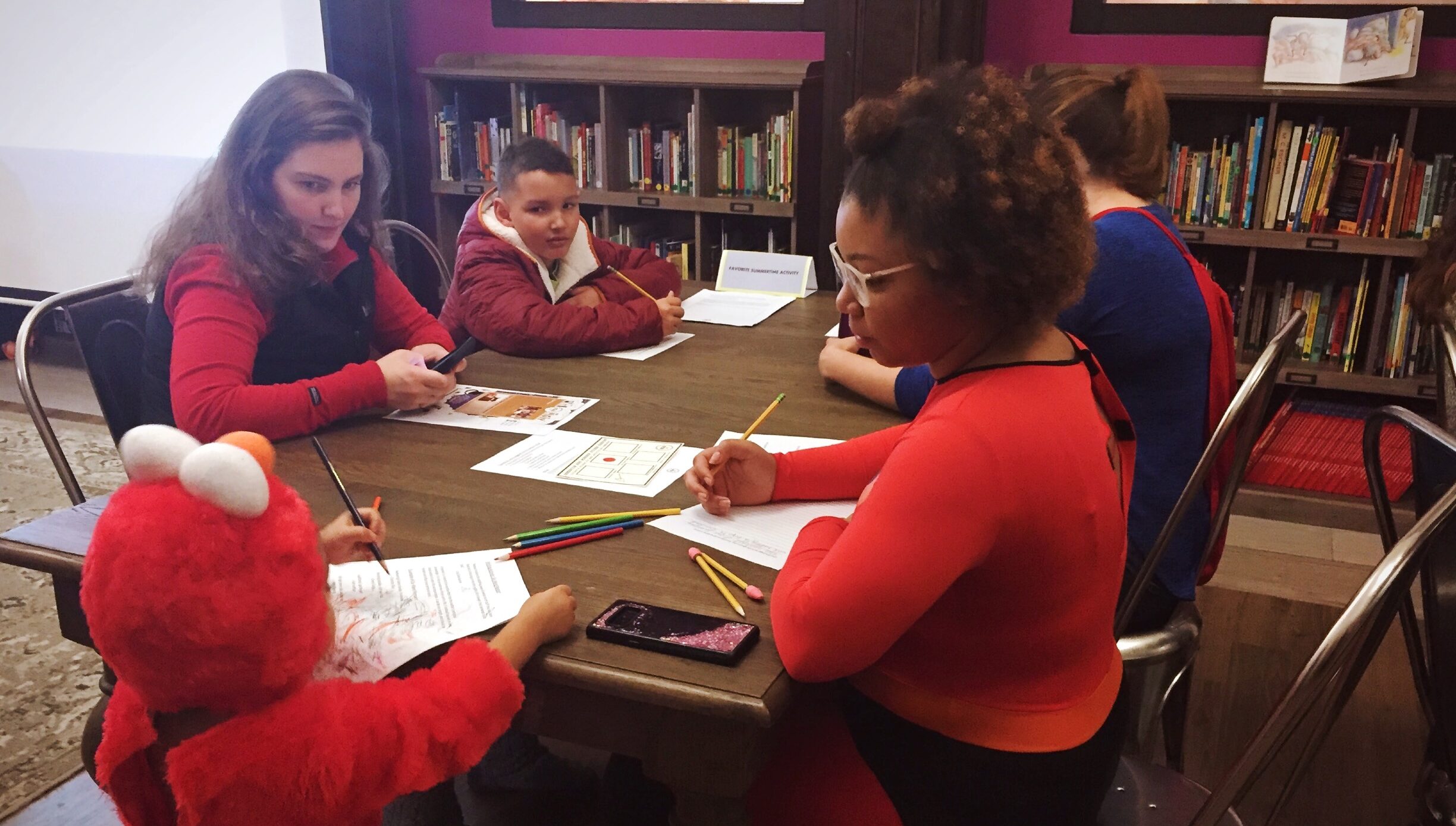 Two volunteers, a boy, and a girl in an Elmo costume sit at a table and write.