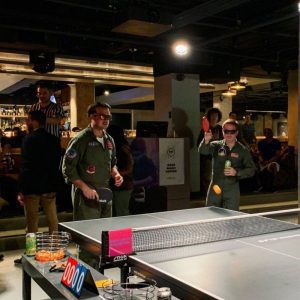 Two men in sunglasses wearing jumpsuits play ping pong